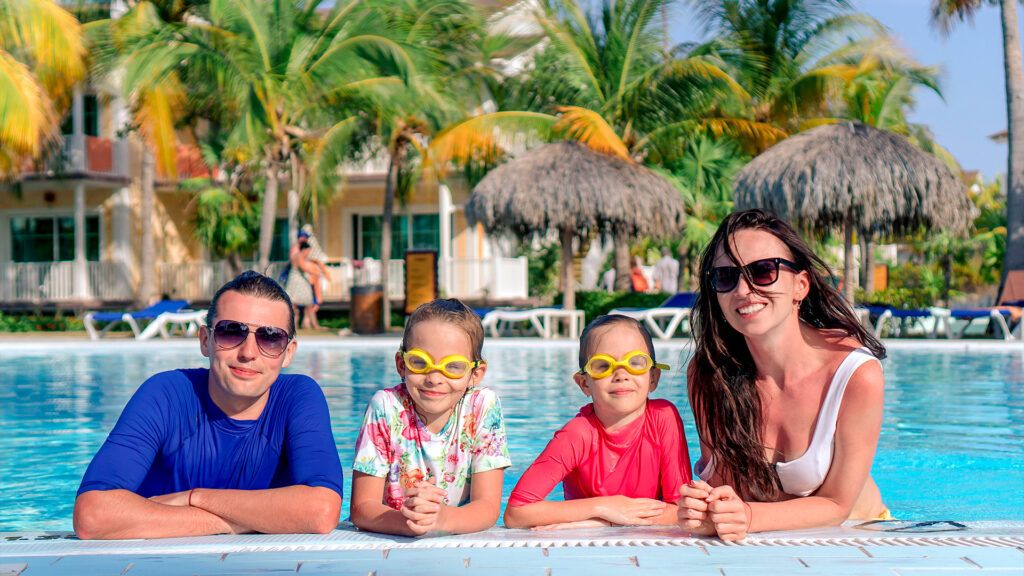 Family of four enjoying the pool together, with children wearing swimming goggles and sunshades at a tropical resort.