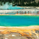 A wide-angle view of the Grand Prismatic Spring in Yellowstone National Park, with its colorful bands of orange and blue, and visitors walking on the boardwalk.