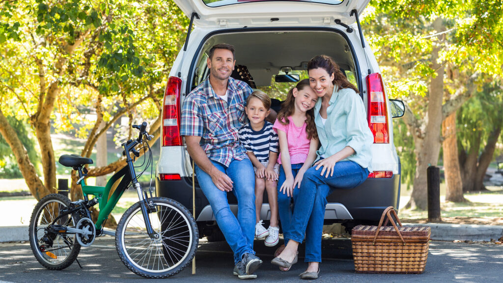A family of four smiling and sitting on the back of their car during a road trip, with a bicycle parked nearby and a picnic basket next to them.