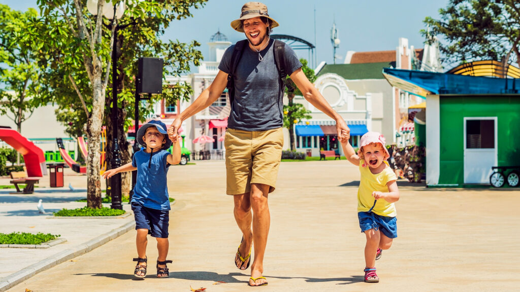 A father walking with his two children in a theme park, holding hands and smiling, ready for a day of fun and adventure.