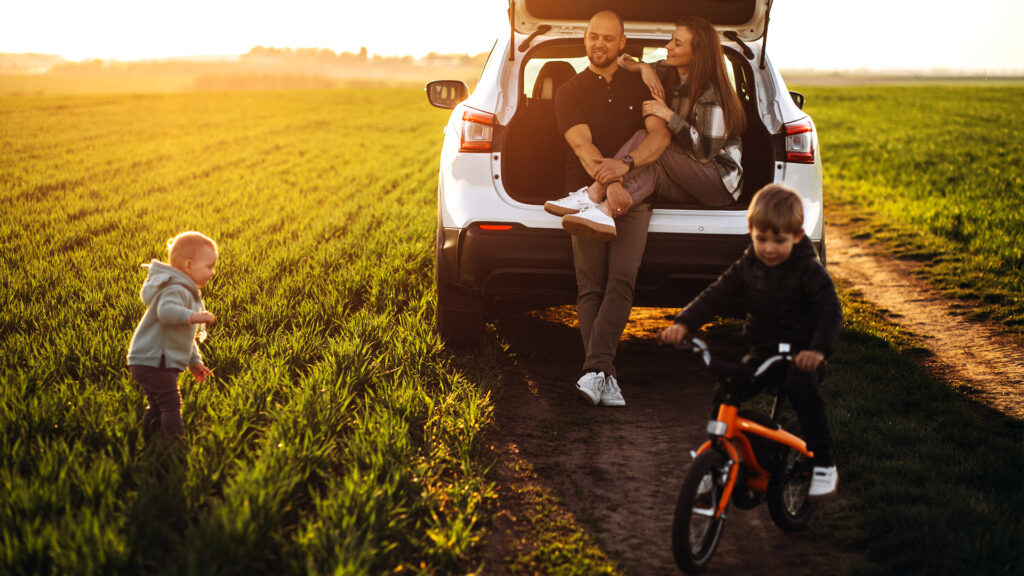 A family enjoying a sunset during their road trip, sitting in the back of their car while a child rides a bike in front of them, surrounded by a field.