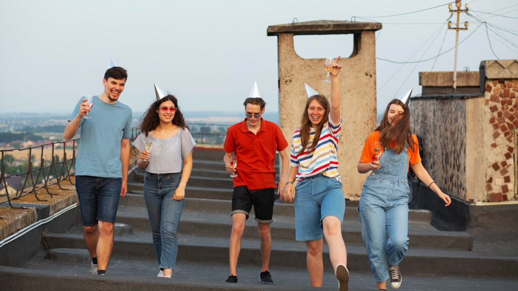Group of five friends celebrating on a rooftop, wearing party hats and holding drinks, enjoying the evening.