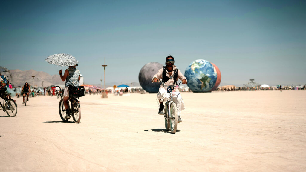 A cyclist rides across the desert landscape at Burning Man, with large art installations in the background.
