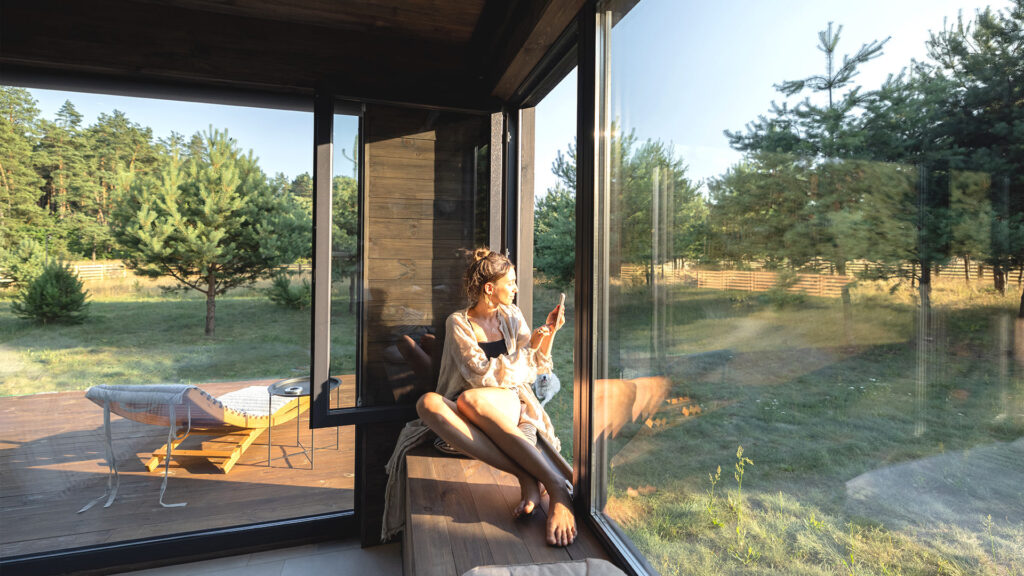 Woman sitting by a large window in a forest cabin, enjoying the serene view, a growing trend in summer travel destinations.