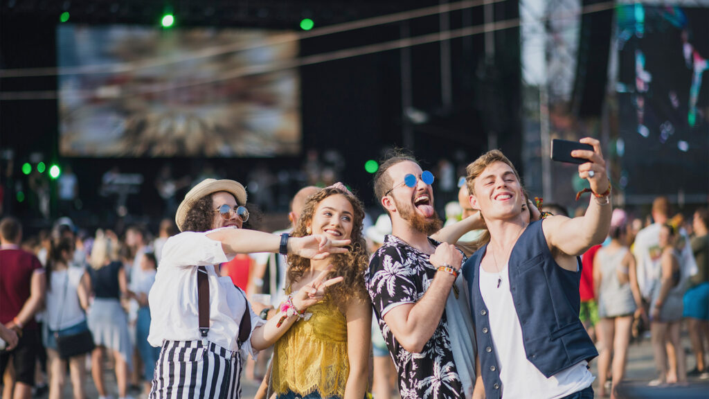 Group of four friends taking a selfie at a music festival with the stage in the background.
