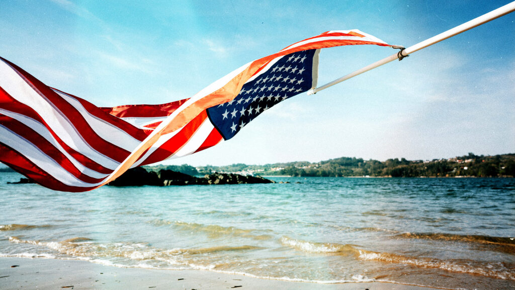 American flag waving on a beach with clear skies and waves in the background.