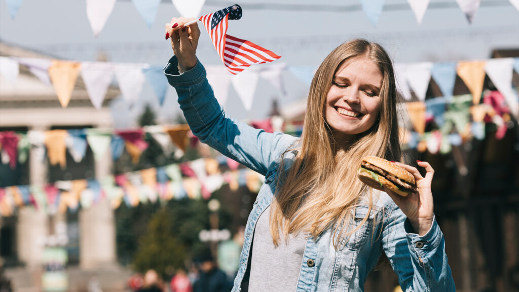 Smiling woman holding an American flag and a burger at a summer festival with colorful bunting decorations.