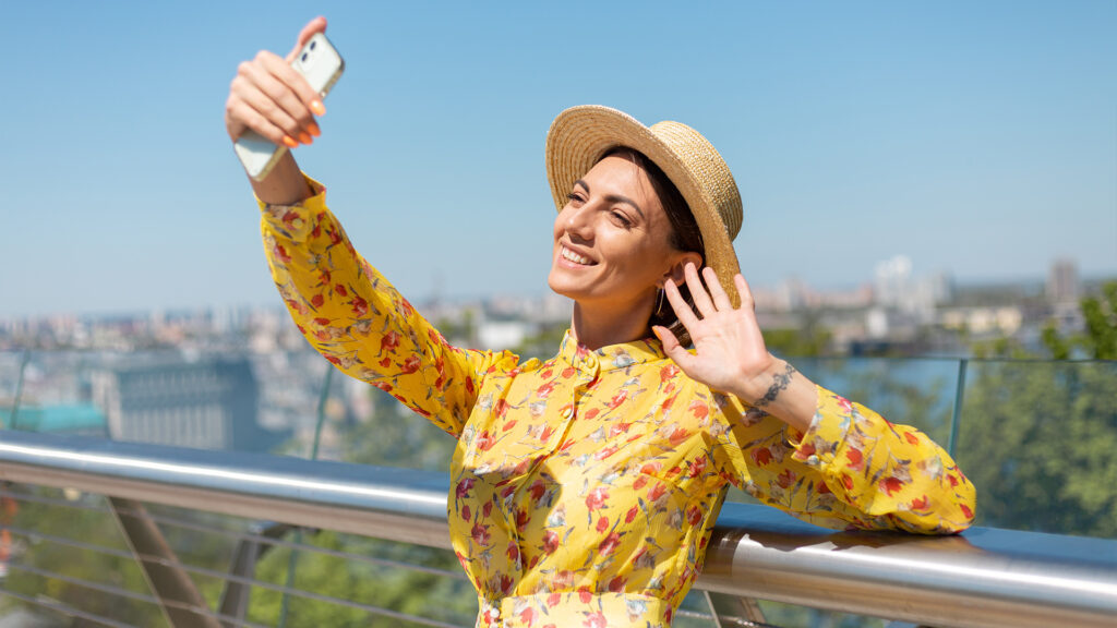 A woman taking a selfie with a sunny cityscape behind her, capturing the essence of summer travel trends.