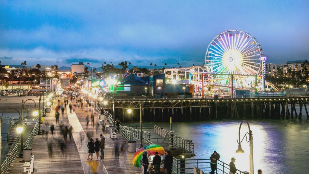 A colorful, illuminated Santa Monica Pier during a summer evening with a bustling crowd and Ferris wheel.