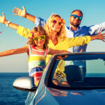 A family having a great time on a road trip, standing on the edge of a convertible car near the ocean, with bright sunglasses and a colorful pinwheel.