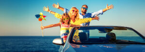 A family having a great time on a road trip, standing on the edge of a convertible car near the ocean, with bright sunglasses and a colorful pinwheel.