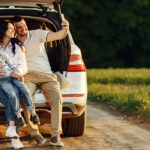 A family of three taking a selfie while sitting on the back of their car parked on a scenic road, enjoying a road trip.
