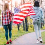Two women walking through a forest path holding the American flag, surrounded by tall trees and greenery.