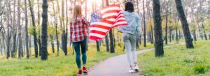 Two women walking through a forest path holding the American flag, surrounded by tall trees and greenery.